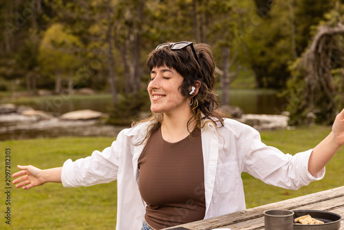 Woman with curly hair sits in a park. Her arm rests on a wooden railing. Sunglasses cover her eyes. Lush green grass and trees surround a small pond. Her calm expression shows peaceful thought
