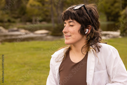 Woman with curly hair sits in a park. Her arm rests on a wooden railing. Sunglasses cover her eyes. Lush green grass and trees surround a small pond. Her calm expression shows peaceful thought