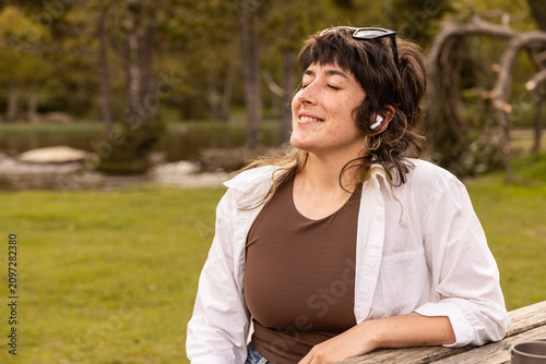 Woman with curly hair sits in a park. Her arm rests on a wooden railing. Sunglasses cover her eyes. Lush green grass and trees surround a small pond. Her calm expression shows peaceful thought