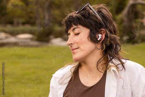Woman with curly hair sits in a park. Her arm rests on a wooden railing. Sunglasses cover her eyes. Lush green grass and trees surround a small pond. Her calm expression shows peaceful thought