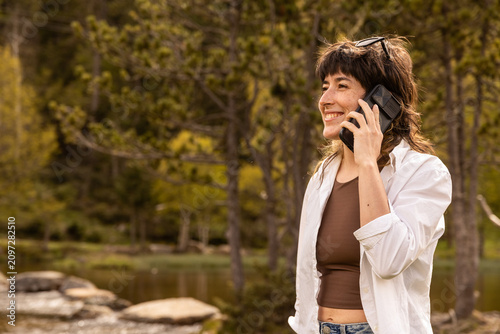 Woman smiles while taking a selfie at a wooden picnic table. She wears a white cardigan over a brown top. Her arms stretch wide as she captures the moment. Coffee mug and book rest on the bench