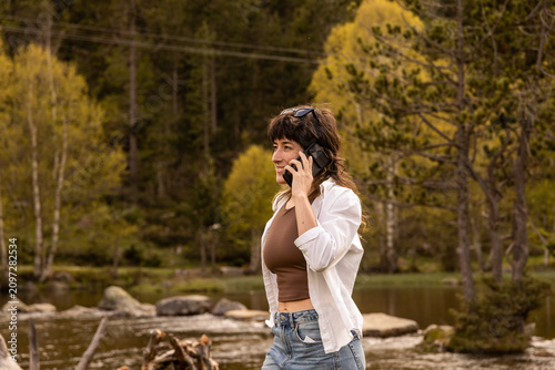 Woman smiles while taking a selfie at a wooden picnic table. She wears a white cardigan over a brown top. Her arms stretch wide as she captures the moment. Coffee mug and book rest on the bench