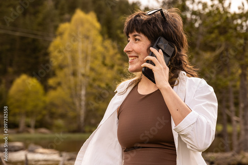Woman smiles while taking a selfie at a wooden picnic table. She wears a white cardigan over a brown top. Her arms stretch wide as she captures the moment. Coffee mug and book rest on the bench