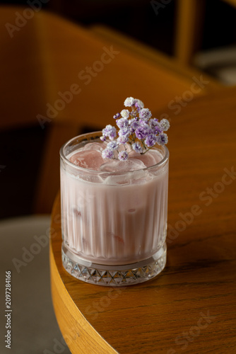 A drink with ice rests on a wooden table. Small flowers are placed on top of the drink. The setting shows warm colors and a cozy atmosphere