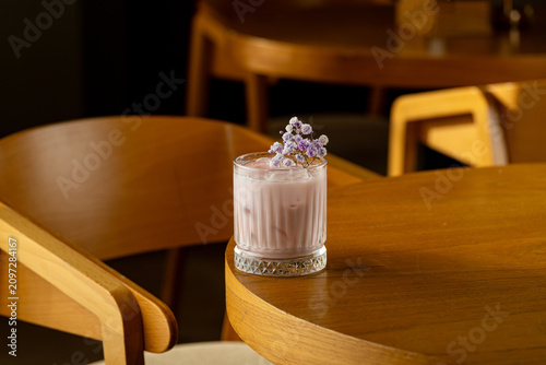 A drink rests on a brown wooden table in a cafe. A small flower decorates the drink. The space has warm tones and simple furniture