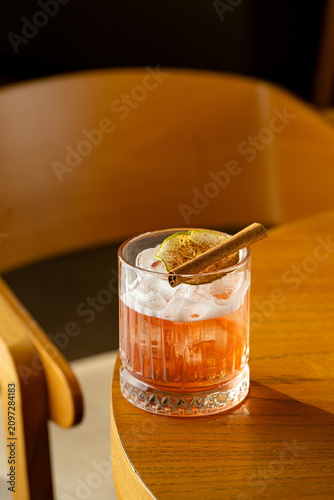 A cocktail glass sits on a wooden table. The drink has ice and a slice of citrus on top. Nearby is a cinnamon stick, creating a warm atmosphere. Sunlight shines in