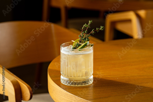 A glass filled with a refreshing drink sits on a wooden table. It is garnished with lemon and fresh herbs. The background shows a simple and inviting space