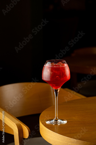 A glass filled with a bright red drink sits on a wooden table. The setting is a dimly lit cafe with a nearby chair. The drink has ice cubes and looks refreshing