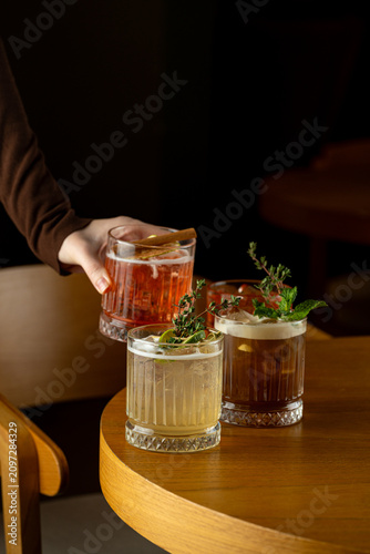 A hand holds a glass filled with a drink, while two other glasses sit on the wooden table. The setting is dimly lit, adding to the atmosphere of the moment