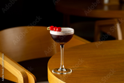 A cocktail with red cherries sits on a wooden table in a bar. The scene shows a relaxed setting where people can enjoy drinks late in the evening