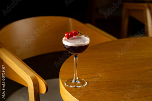A cocktail sits on a wooden table in a bar. The drink has a layer of foam and is topped with a few cherries. The setting is dark and cozy, with wooden furniture