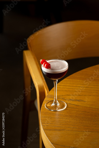 A cocktail sits on a wooden table next to an empty chair. The drink features a garnish on the rim. The setting shows a bar atmosphere during the evening