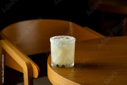 A cocktail sits on a wooden table in a bar. The drink has a salt rim and is clear. The setting is dimly lit and creates a relaxed atmosphere for evening socializing