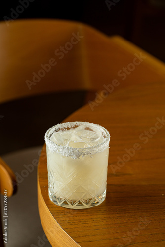 A glass filled with a mixed drink is placed on a wooden table. The drink has ice and a salt rim. The setting is inside a bar with warm lighting