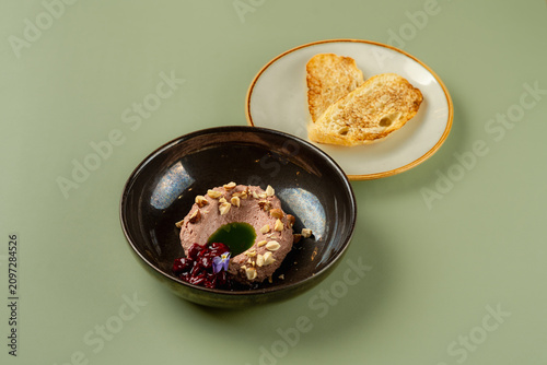 A dessert is placed in a dark bowl. It has nuts and fruit on top. Slices of toasted bread are on a plate next to the bowl. The scene is set on a green table surface