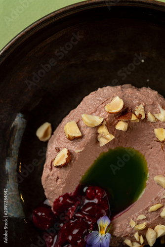 A dessert is placed in a dark bowl with a smooth texture. The dish has nuts, dark sauce, and a green sauce on top. The setting is simple, showing focus on the food