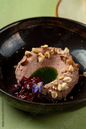 A round dessert with a hole in the center is placed in a black bowl. The dish includes sauce on one side and is topped with nuts. The setting is on a smooth green table