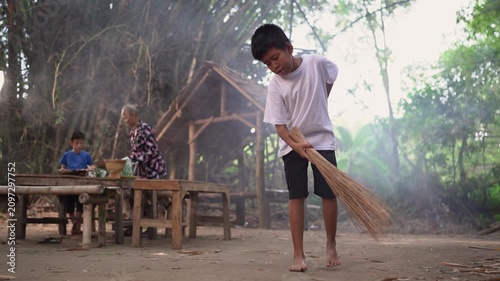 Young boy sweeps dusty ground in foreground beside rustic bamboo shelter, while older woman and seated child remain near wooden table in background