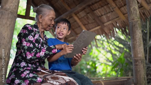 Elderly woman leans beside smiling young boy as both focus on handheld tablet inside rustic open-air bamboo shelter, with woven thatch roof, rough wooden posts, soft daylight, and calm rural