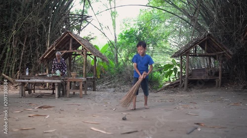Young boy in blue shirt sweeps dusty earth across quiet rural courtyard framed by dense bamboo, rustic thatched shelters, simple table, stools, and lush green foliage in bright daylight