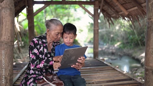 Elderly woman and young boy sit close together on bamboo platform inside open rustic shelter, sharing tablet with focused smiles, soft daylight, green background, and calm village stream beside
