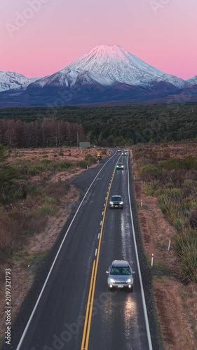 A scenic road winds through the Tongariro National Park in New Zealand, leading towards the majestic Mt Ngauruhoe at sunrise. The golden light illuminates the landscape.