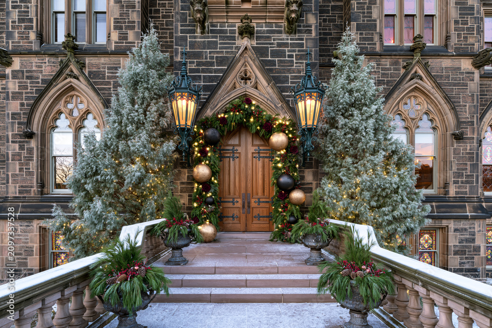 Fototapeta premium Gothic mansion entrance decorated for Christmas with lanterns, evergreen garland, oversized ornaments and frosted trees.