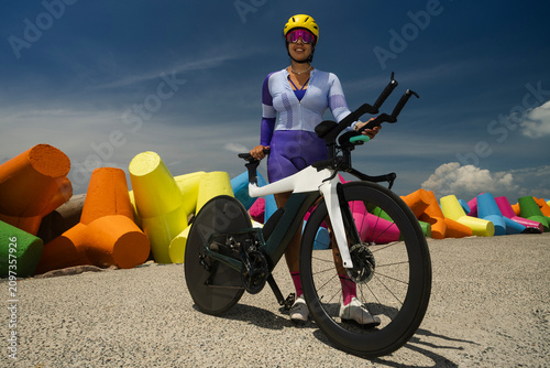 Female triathlete standing with time trial bicycle in colorful coastal setting wearing cycling helmet