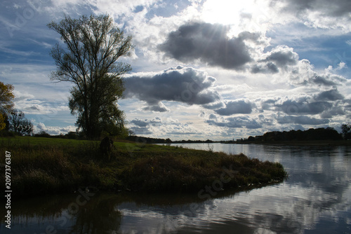 A solitary tree beside a calm river beneath dramatic clouds. A peaceful landscape featuring reflections, natural beauty and a wide open riverside scenery.