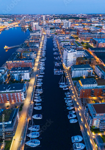 Drone photo high above Malmö harbor at blue hour, summer evening 2024, capturing glowing city lights, calm water reflections, and Scandinavian urban beauty in cinematic aerial perspective.