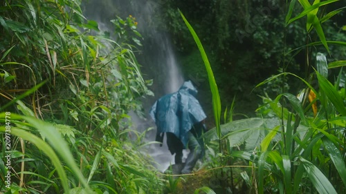 A person wearing a rain poncho walking at a massive waterfall, with water cascading down with great force amidst the inhospitable jungle