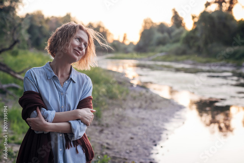 Schilderij op canvas miling woman wrapped in a shawl standing by a peaceful river at sunset, enjoying a quiet moment in nature