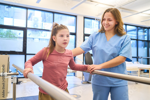 Child physiotherapy session, girl walking with parallel bars, supported by therapist, pediatric rehabilitation exercises for motor skills training, in a bright and modern clinic