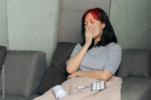 Sick asian woman having severe cough resting on grey sofa bed at home feeling exhausted and expressing painful fever headache while taking prescription medicine pill for illness treatment