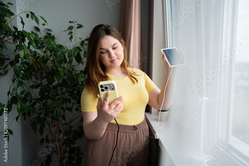 Young woman charging smartphone with portable solar panel by window indoors. Renewable energy sustainable technology and mobile power solution