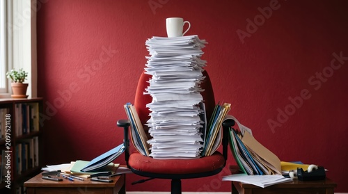 Tall stack of paperwork piled on office chair with folders and scattered documents on desk against red wall, overwhelmed desk clutter suggesting heavy workload and organization challenge.