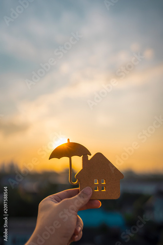 Hand holding a wooden house cut-out with an umbrella silhouette against sunset. Concept of home insurance, financial protection, property safety, and mortgage security for family.