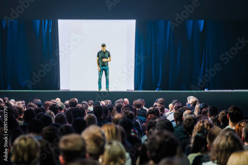 Large audience focused on lone speaker delivering presentation on illuminated stage, dramatic lighting and attentive crowd in conference auditorium.