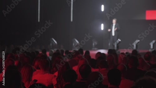 Audience watching keynote speaker on stage at corporate conference. Dramatic red lighting creates moody, focused atmosphere.