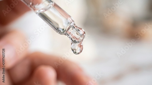 Glass dropper with a clear liquid dripping on a blurred background