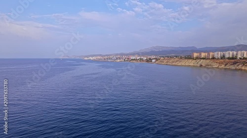 The camera shows a wide view of the coastline with clear blue water and distant buildings. The sun sets, creating a warm light over the sea and land.
