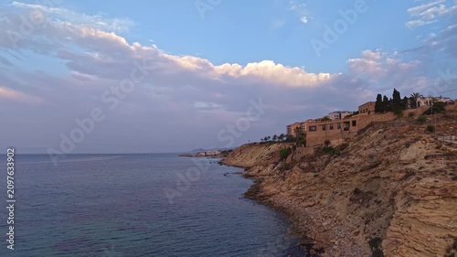 The camera shows a stretch of the coast with buildings above the water. Clouds fill the sky as the sun sets, creating a natural view of the ocean below.