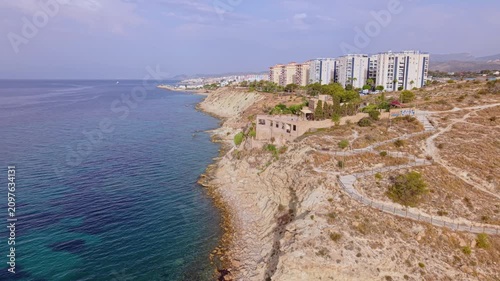 Aerial shots capture buildings along the coast. The landscape features rocky cliffs and blue water. The sun shines down, highlighting the natural beauty of the area.