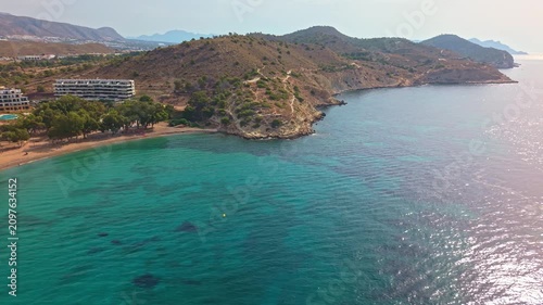 A view shows the beach and clear water near a resort. Sunlight shines on the waves. Visitors walk along the shore. Mountains surround the area in the background.