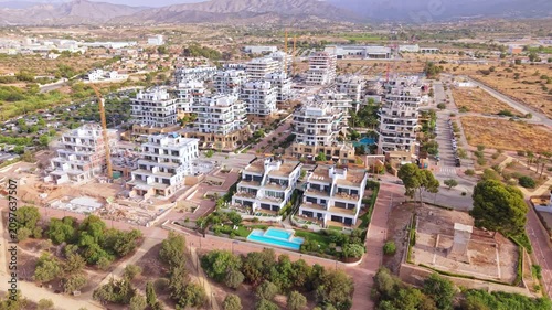Aerial shots show a developing neighborhood with several new buildings and open spaces. Green areas and roads surround the structures. The time is daytime, and the sky is clear.