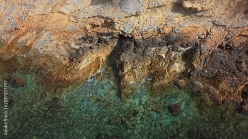 People observe the rocky coastline by the clear water. The sun shines down, and waves gently touch the rocks. It is midday, and the area shows signs of nature's beauty.