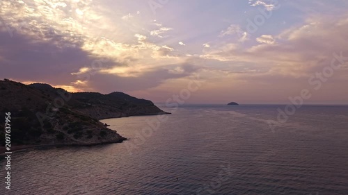 The sun sets over the sea, casting colors in the sky. Waves move gently against the shore. Hills frame the coast while an island is seen in the distance. Evening light creates a soft view.