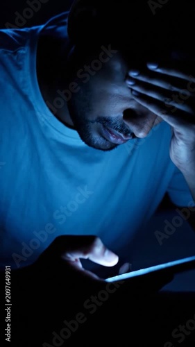 Stressed man using smartphone in dark room with blue light.