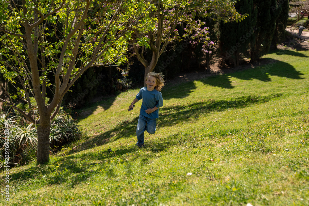 Fototapeta premium Little blond boy running on green hill in sunny park, childhood freedom concept, outdoor activity, joyful movement, spring nature, lifestyle scene, carefree play, landscape background