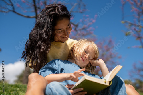 Young mother and little son reading book outdoors in sunny park, child tired and resting, family bonding, education, calm peaceful moment, leisure lifestyle, spring day